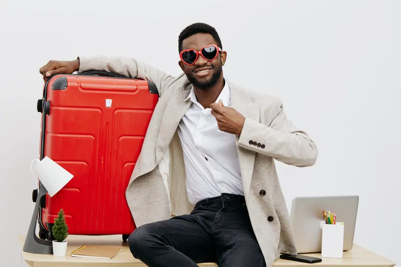 Smiling confident man with dark skin, wearing beige coat and white shirt, sitting at desk with red suitcase, laptop, and stationery, on plain white background, studio setting, travel and business conc