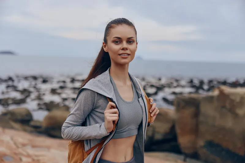 Young athletic woman in activewear on a rocky beach, smiling confidently with a backpack, promoting healthy lifestyle and outdoor fitness activities