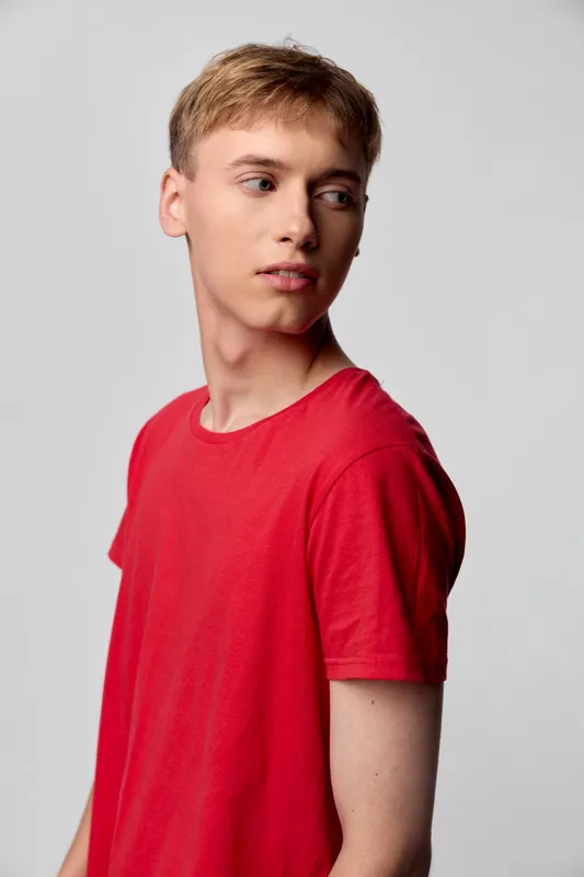 Young man in a red shirt posing in a clean studio, looking away from camera, calm expression, simple background, casual fashion scene, clear focus on clothing and posture.