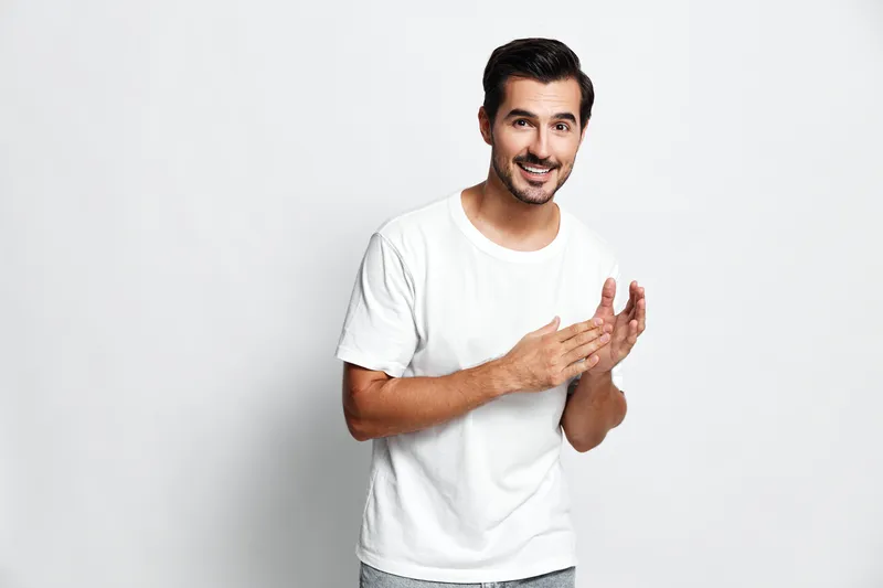 Smiling man in casual white t-shirt, looking at camera with cheerful expression, hands clasped together, isolated on plain light gray background. People lifestyle concept