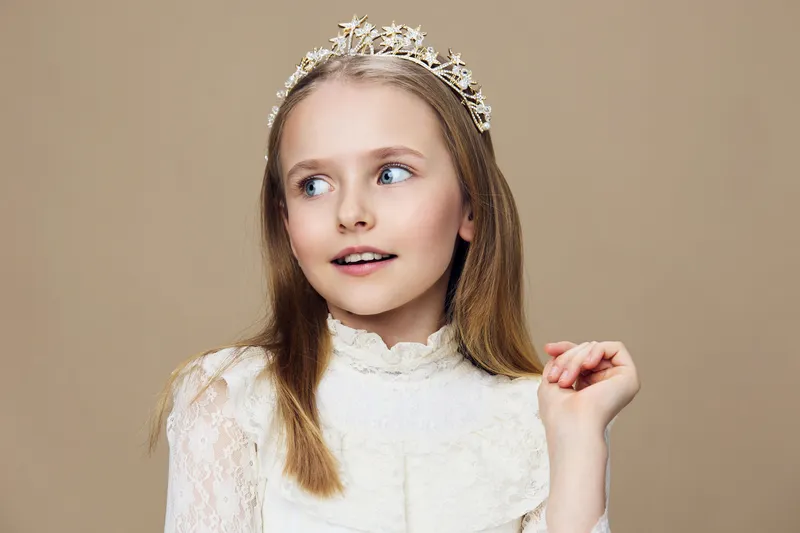 Young girl in elegant attire with a tiara, displaying a charming smile against a soft beige background, perfect for fashion or beauty themes