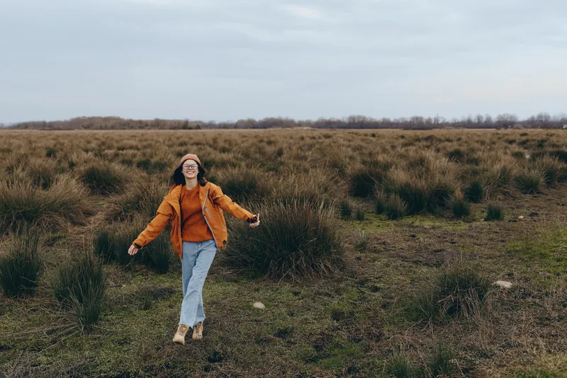 Woman lifestyle walking outdoors in a grassy field, orange jacket, light jeans, and a hat, relaxed pose, nature landscape under cloudy sky, soft natural light, wide horizon, calm mood