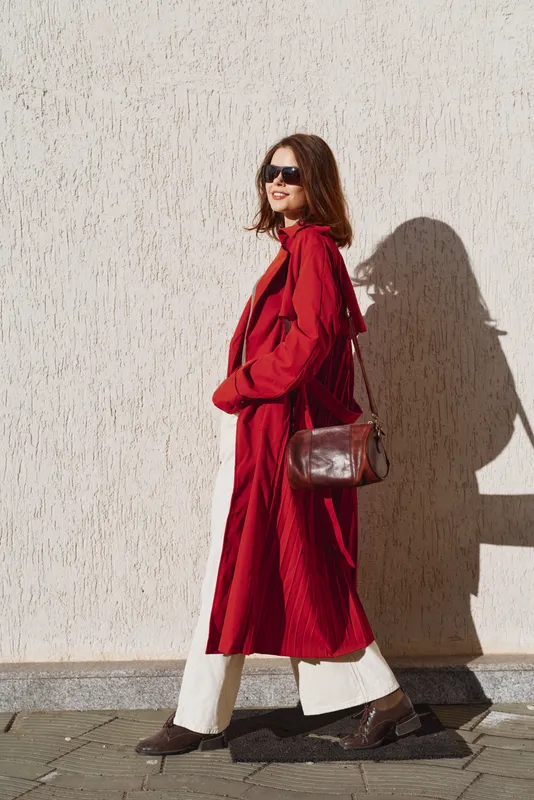 A stylish woman in a red coat walks along a sunlit wall, wearing sunglasses and a brown leather bag, white wide pants peeking from under the coat, exuding confident urban flair.