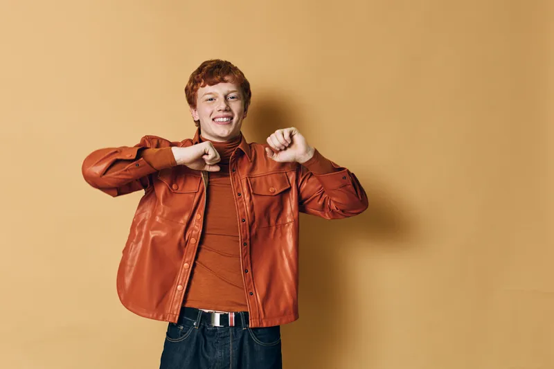 Happy teenage boy in orange jacket dancing and smiling against beige background. Casual outfit with jeans and cheerful mood for youth lifestyle, fun and freedom concept.