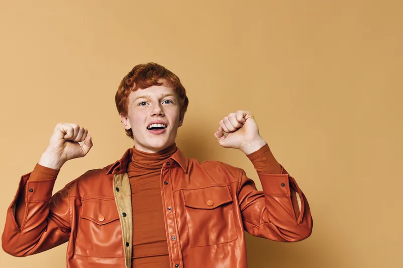 Young man wearing orange shirt and jacket with red hair celebrating success with raised fists and happy face standing against orange background in studio.