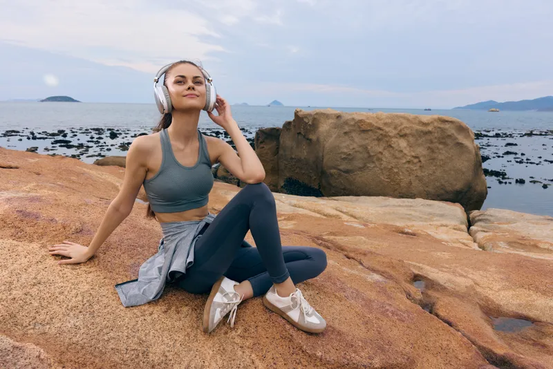 Young woman listening to music on wireless headphones while sitting on rocky beach during a sunny day, wearing sporty outfit and enjoying the calm sea view