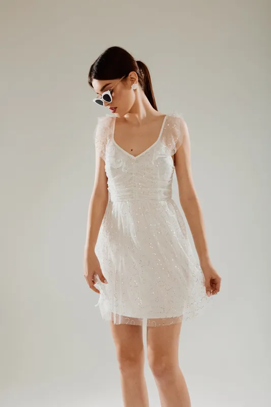Young woman in a white lace dress wearing sunglasses looks down thoughtfully. Studio shot on a neutral background. Concept of woman beauty fashion and elegance.