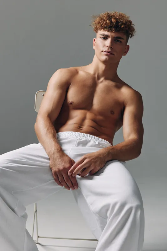 Young muscular man with curly hair posing shirtless in white pants sitting on chair against gray background, fitness model with defined abs and confident expression.