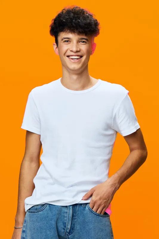 smiling young man wearing white t-shirt and blue jeans stands against bright orange background looking happy and casual with curly hair and relaxed posture