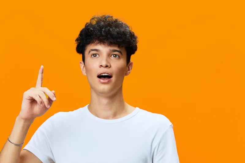 young man with curly hair wearing white t-shirt pointing upwards with surprised expression on bright orange background, casual style and lively emotion
