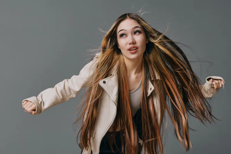 Young woman with long straight hair wearing light jacket poses with hair flying on gray background. Beauty model portrait with casual style and confident expression in studio lighting.