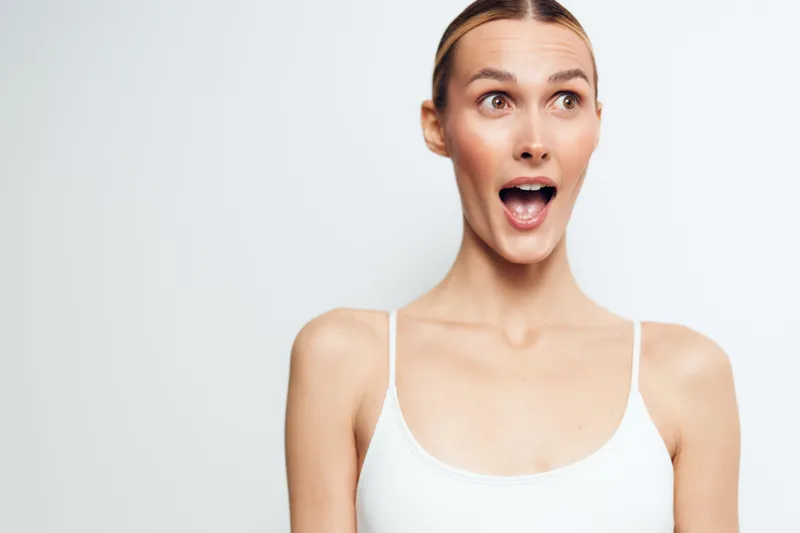 Surprised young woman with blonde hair looking away with open mouth on white background. Casual outfit with thin straps. Concept of emotion, reaction, and expression.