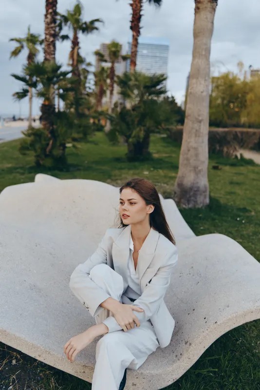 Confident woman in a business suit sitting on a sculptural bench surrounded by palm trees, exuding style and professionalism in an urban setting.