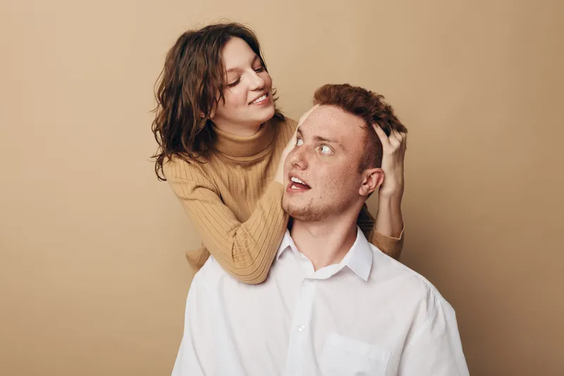 Young couple in casual clothes share a playful moment, woman smiling and touching man’s hair. Beige background creates warm, intimate atmosphere. Concept of love, relationship, and happiness.