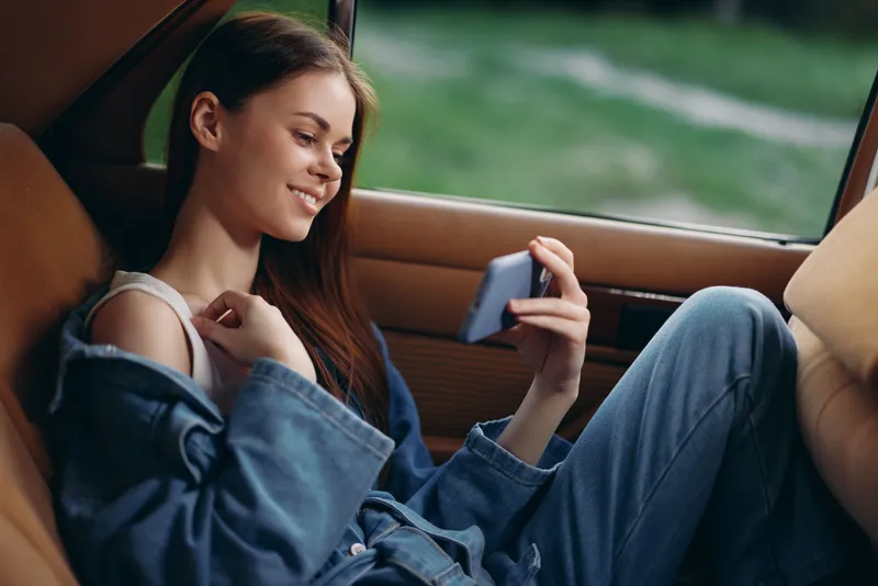 Young woman enjoying her time in a vintage car, smiling while using her smartphone, showcasing casual denim fashion and a relaxed atmosphere.