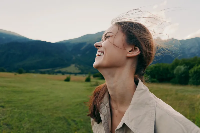Young woman in a light shirt smiles with windblown hair in a sunlit meadow as green hills and distant mountains provide a serene natural backdrop.