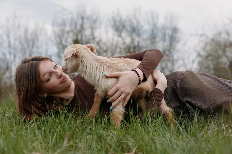 Relaxed woman outdoors, cuddling a curious goat kid in a sunny grassy field, joyful moment, soft breeze, natural colors, serene rural scene, warm connection between human and animal