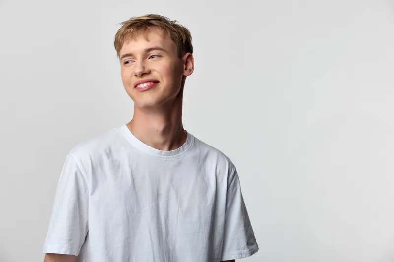 Smiling man in a white t shirt stands against a neutral studio backdrop, exuding casual style and approachable confidence in a clean, modern portrait setup