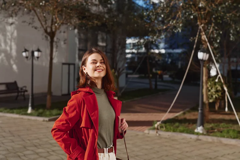 A confident young woman in a red coat smiles outdoors on a sunny day, enjoying the autumn weather in an urban park with trees and paved walkways.