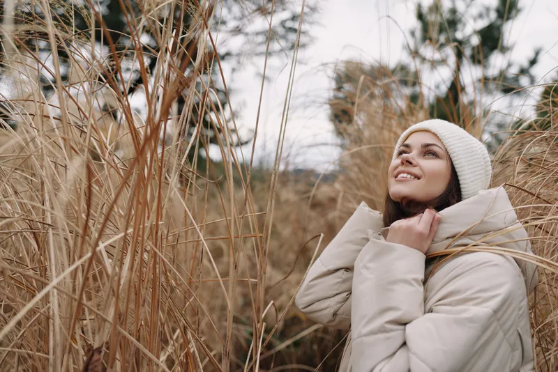Woman in a warm coat and beanie standing among tall dried grass, smiling softly in a crisp outdoor scene. Cozy winter mood, nature backdrop, calm expression, gentle breeze and light.