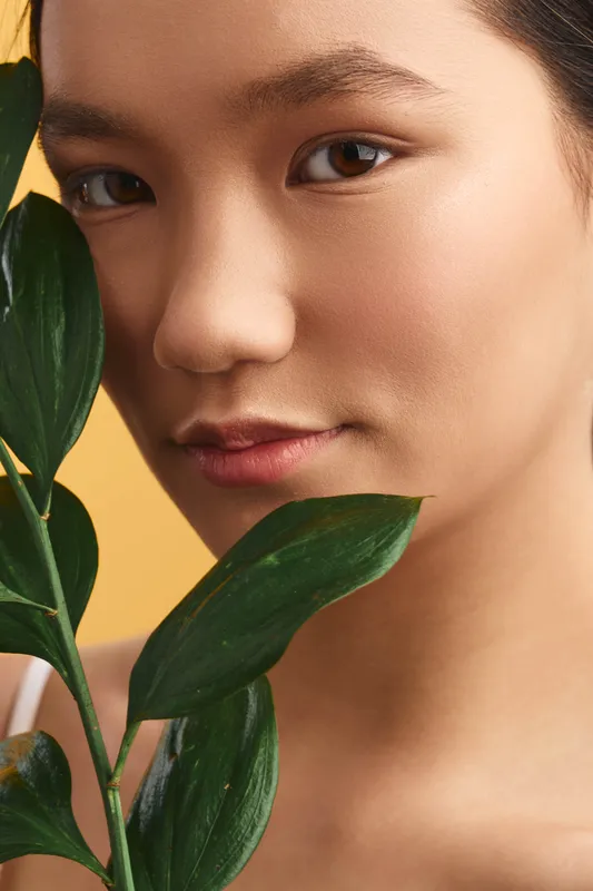 Asian woman with soft makeup gazes through green leaves in a closeup portrait, conveying calm elegance and natural beauty with a warm neutral backdrop.