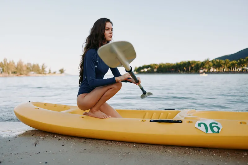 A young woman kneels on a bright yellow kayak at the water's edge, gripping a paddle as soft waves lap the shore under warm sunlight.