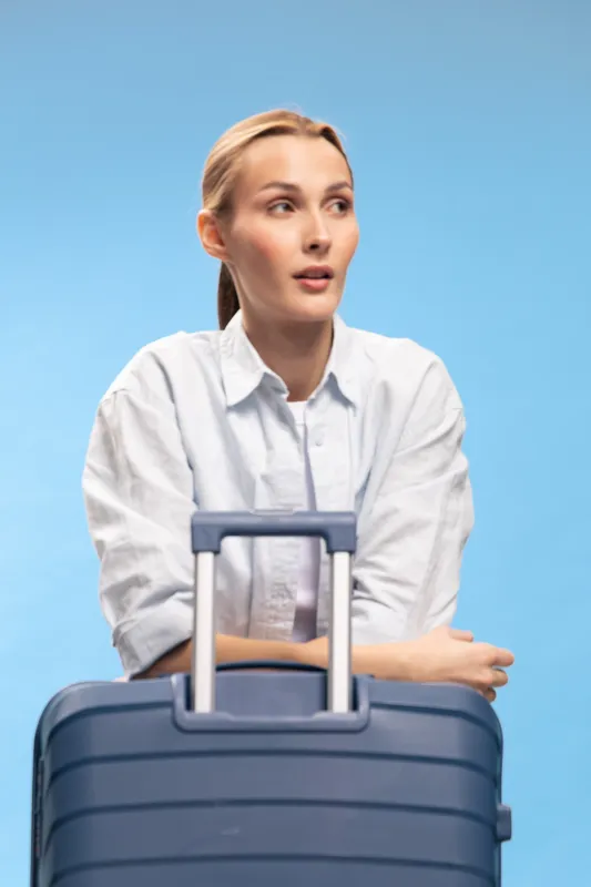 Young woman in white shirt stands with blue suitcase on light blue background. She looks away thoughtfully. Concept for travel, waiting, and journey preparation.