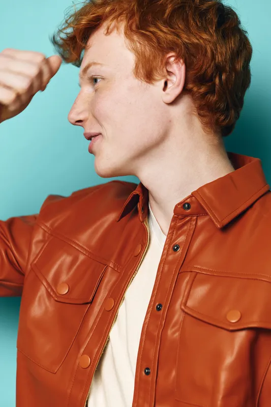 Young man with red hair in orange leather jacket showing strength gesture with fist, side profile on blue background, casual style and confident expression, studio portrait.