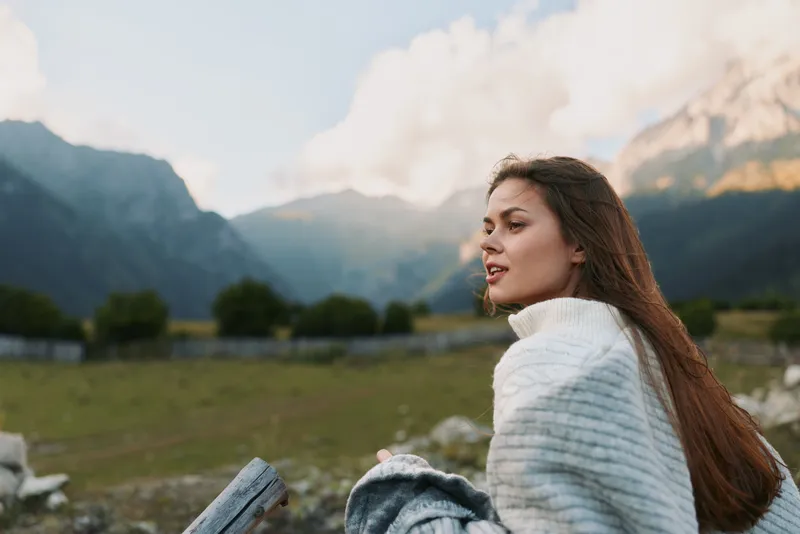 Young woman wearing a white knit sweater sits in a wide valley with distant mountains, looking away with a calm, content expression in a serene outdoor scene.