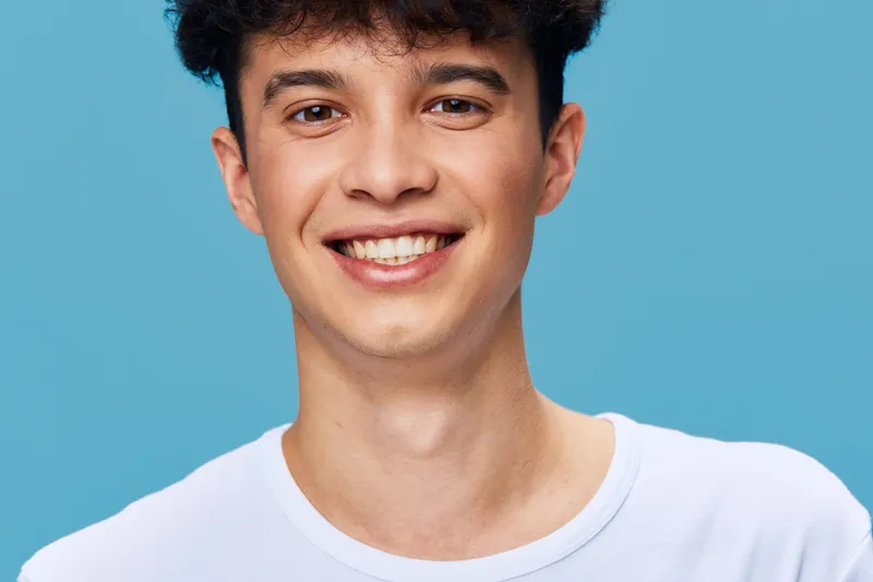 smiling young man wearing a white t-shirt with curly dark hair stands against solid blue background, expressing happiness and youthful energy in casual style.