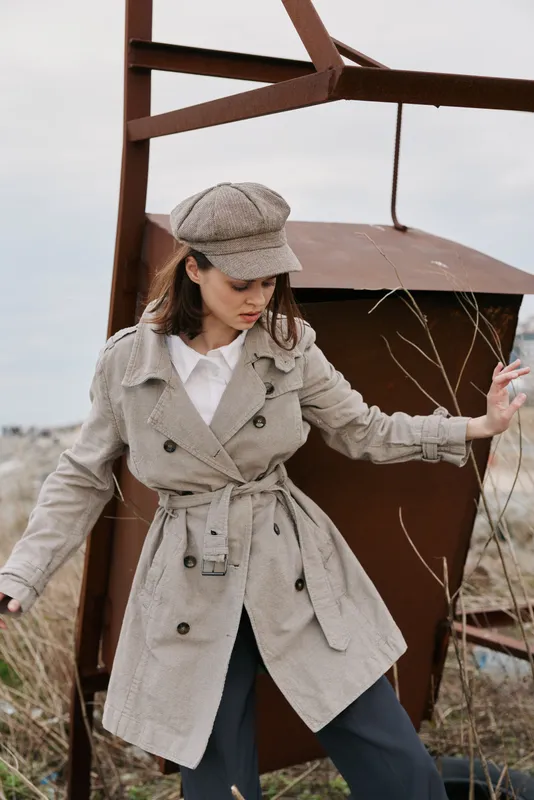 Young woman in beige trench coat and matching newsboy cap poses outdoors near rusty metal structure. Overcast sky and dry grass create calm, muted autumn fashion scene with vintage vibes.