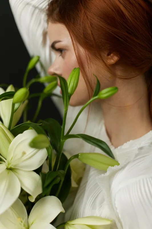 Young woman with red hair holding white lilies, embodying beauty and serenity against a dark background, evoking springtime freshness and elegance