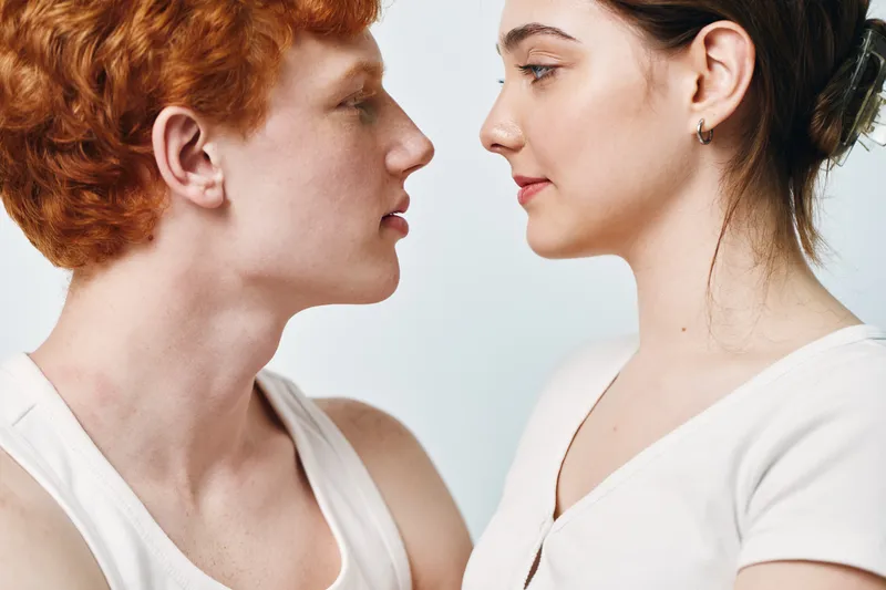 Young man and woman face to face looking at each other closeup in studio with neutral background. Intimate moment, red hair, earrings, casual clothing, profile view of two people.
