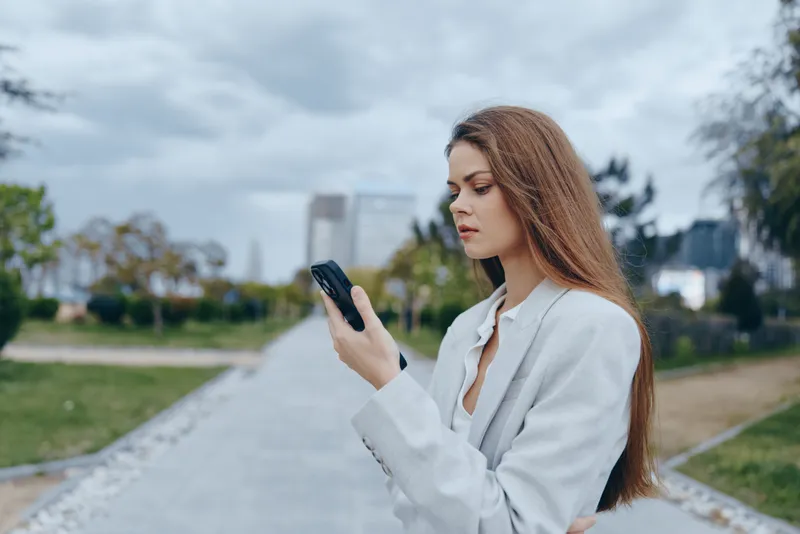 Young woman in light blazer outdoors using smartphone with thoughtful expression in urban park setting autumn season business concept