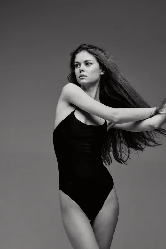 Black and white portrait of a slender young woman in a black swimsuit with flowing hair and serious expression.