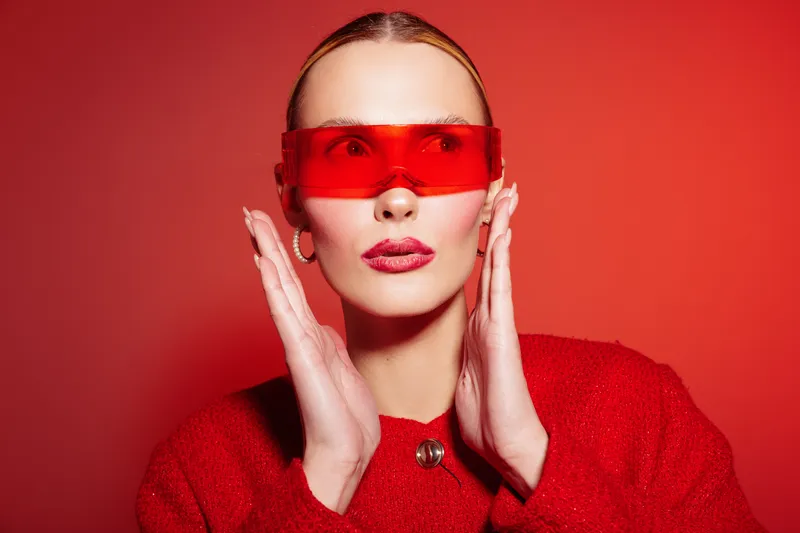 Stylish woman in red sweater and red translucent glasses poses against red background. Fashion portrait with bold makeup and modern accessories. Concept of style and confidence.