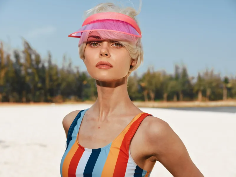Portrait of a stylish woman on a sunny beach wearing a colorful striped swimsuit and pink visor, posing confidently with wind in her hair and bright summer light.