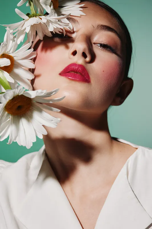 Close-up portrait of a fashionable woman with bright red lipstick and flowers next to face on a green background. Creative beauty concept capturing elegance and style.