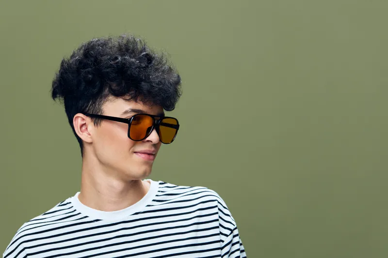 young man with sunglasses wearing a striped shirt, curly hair, casual style, relaxed expression, green background, modern fashion portrait