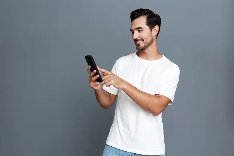 Full body smiling happy cheerful young man wearing white t-shirt and blue jeans standing against gray background, using mobile cell phone with joy. Lifestyle concept.