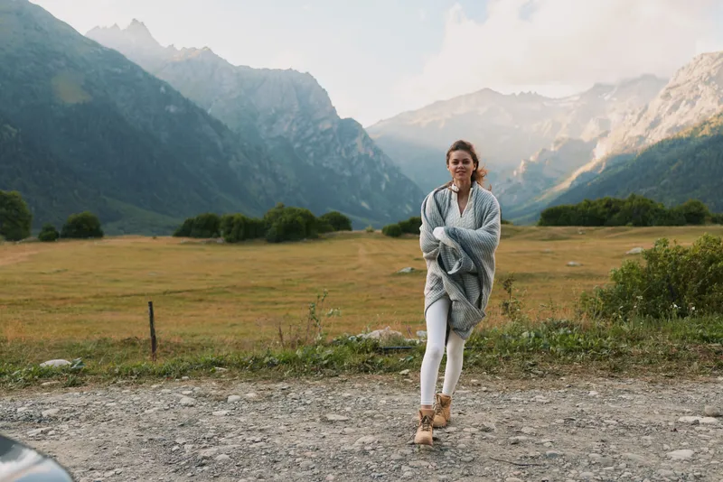 Cheerful woman outdoors, walking through a grassy field with distant mountains, cozy shawl, relaxed mood, natural colors, sunlit landscape, fresh air