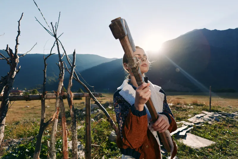 woman with axe in countryside near wooden fence and mountains with sun flare, outdoor rural scene of rustic woodcutting and manual work, portrait of determined person preparing firewood