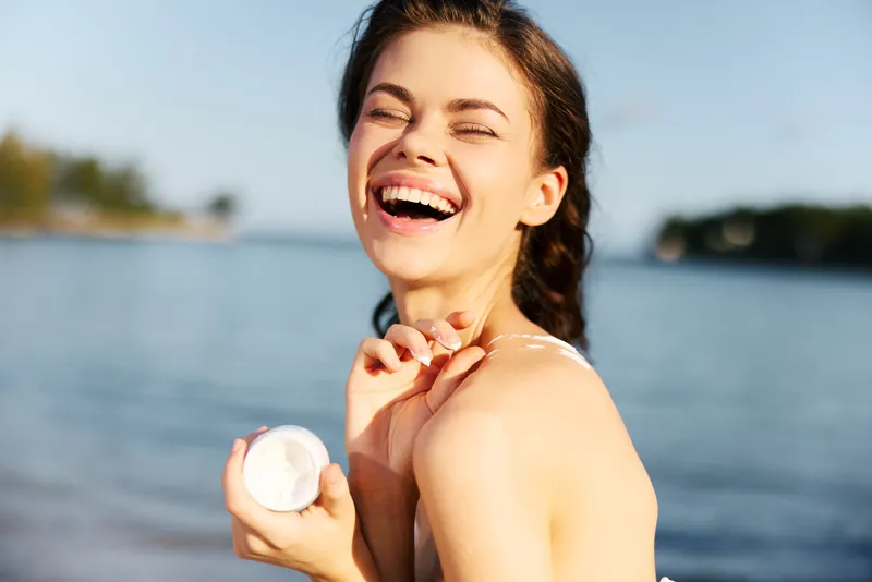 Bright joyful portrait of a smiling woman at the beach, applying sunscreen from a small container while sunlight glints off the water and a warm breeze lifts her hair.