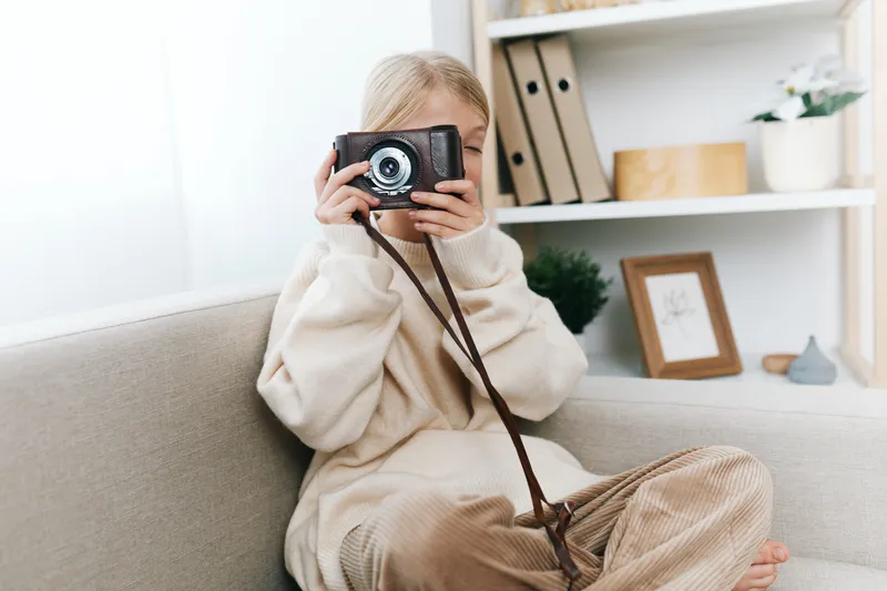Young photographer sits on a cozy sofa, holding a camera up to eye level, soft natural light, warm sweater, relaxed pose, creative lifestyle moment captured at home.