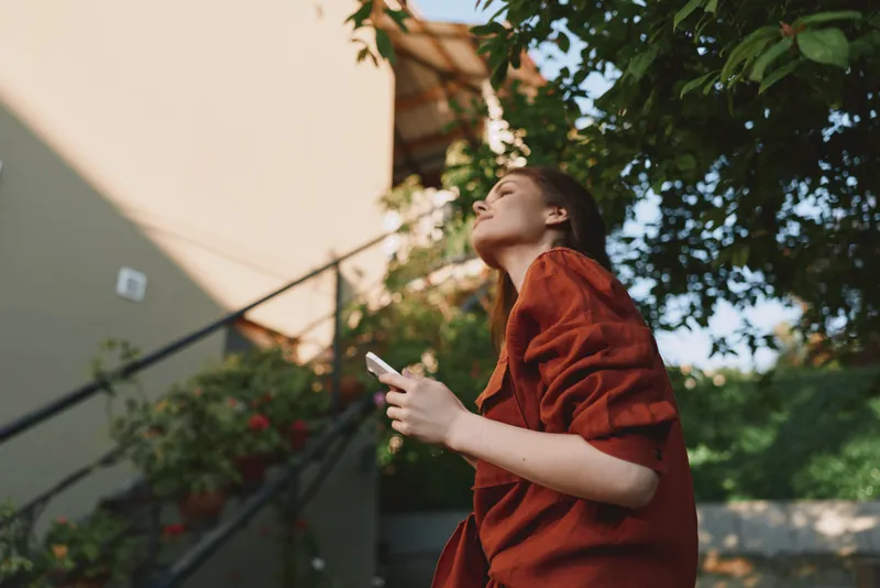 Young woman in a red dress stands outdoors looking upward with a calm expression, sunlight on her face, surrounded by greenery and a pale building in the background