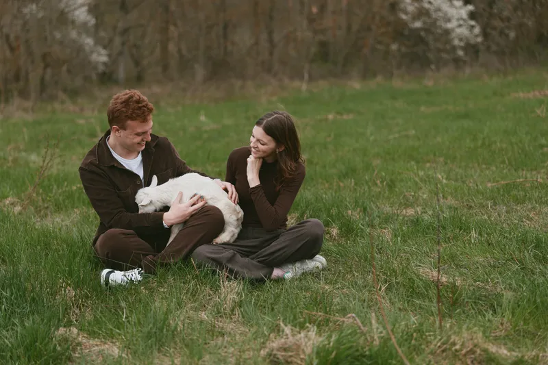 Couple sits on a grassy field outdoors, gentle interaction with a goat kid, smiling and relaxed, natural lifestyle moment captured in soft evening light, in a tranquil park setting