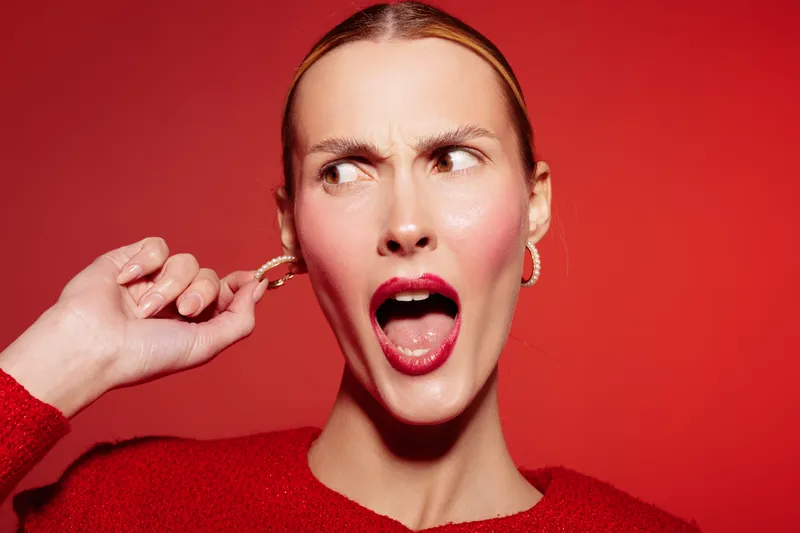 Surprised woman with red lipstick and earrings covers ear with hand, looking sideways on red background. Concept of shock, reaction, and emotional expression in studio lighting.