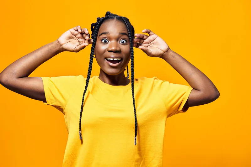 Joyful Black girl with braids in a yellow t shirt expressing excitement against a vibrant yellow background, perfect for concepts of happiness and positivity