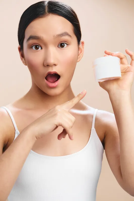 Asian woman holds a white cosmetic jar in a bright studio, smiling with surprise while pointing to the product, suggesting curiosity and fresh skincare allure.