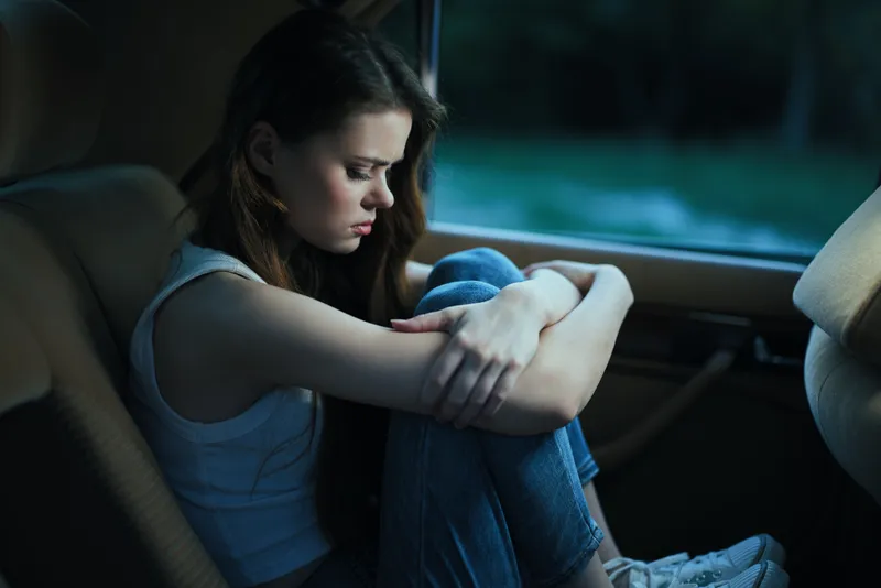 A pensive young woman sits in a car, dressed in casual attire, conveying emotions of sadness and introspection with her arms wrapped around her knees.